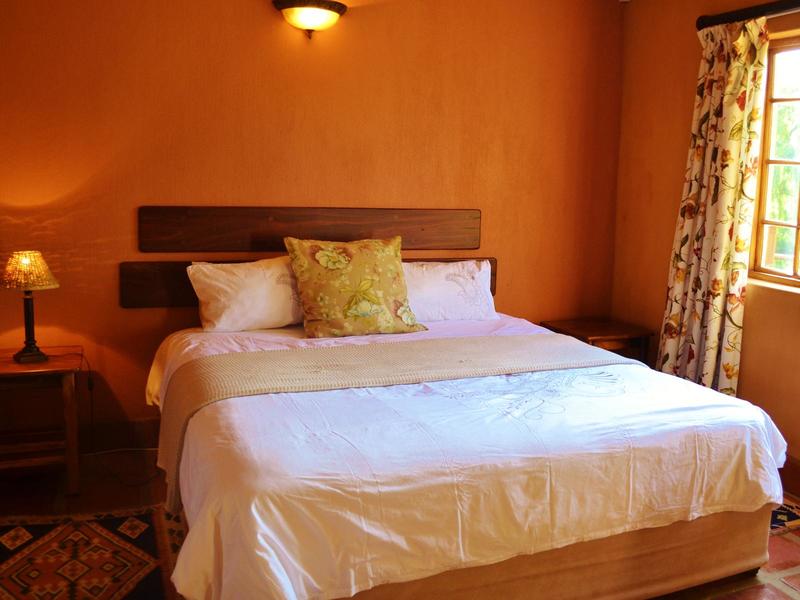 A spacious bedroom featuring a king-size bed with a light-colored duvet and decorative pillows. Wooden bedside tables are positioned on either side, and natural light filters through a window adorned with patterned curtains. The warm orange walls create a cozy ambiance.