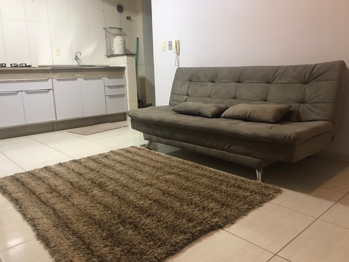 A neutral-colored sofa with soft cushions is positioned in front of a simple kitchenette, complemented by a textured area rug on the tiled floor. The space is well-lit, with bright walls enhancing the open layout.