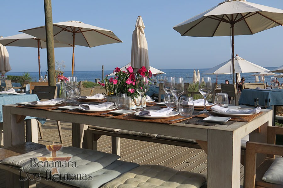 A dining table is set for multiple guests, featuring glassware and neatly arranged dishes. The scene is enhanced by vibrant flowers in the center and large umbrellas providing shade. In the background, the beach and ocean are visible under a clear blue sky.