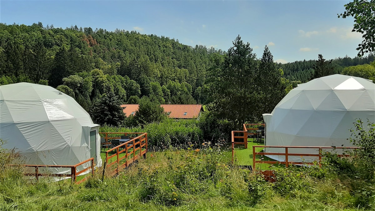 Two luxurious igloos are nestled amid greenery, surrounded by forested hills. Each geodesic structure features a white exterior and is elevated on wooden decks. A glimpse of the resort buildings can be seen in the background, offering a serene atmosphere.