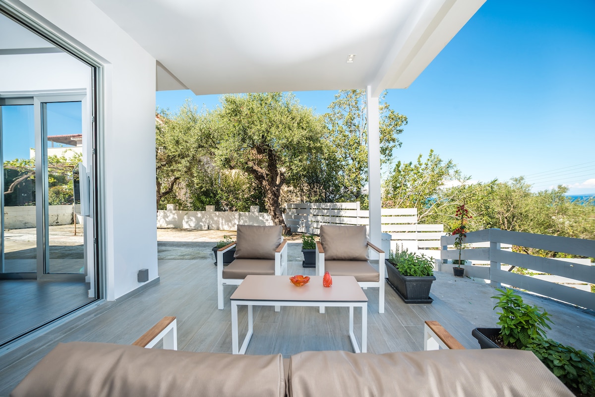An outdoor seating area is visible, featuring two light brown chairs and a matching table. Lush greenery surrounds the space, and a glimpse of the water can be seen in the background. Natural light brightens the area, enhancing the relaxed atmosphere.