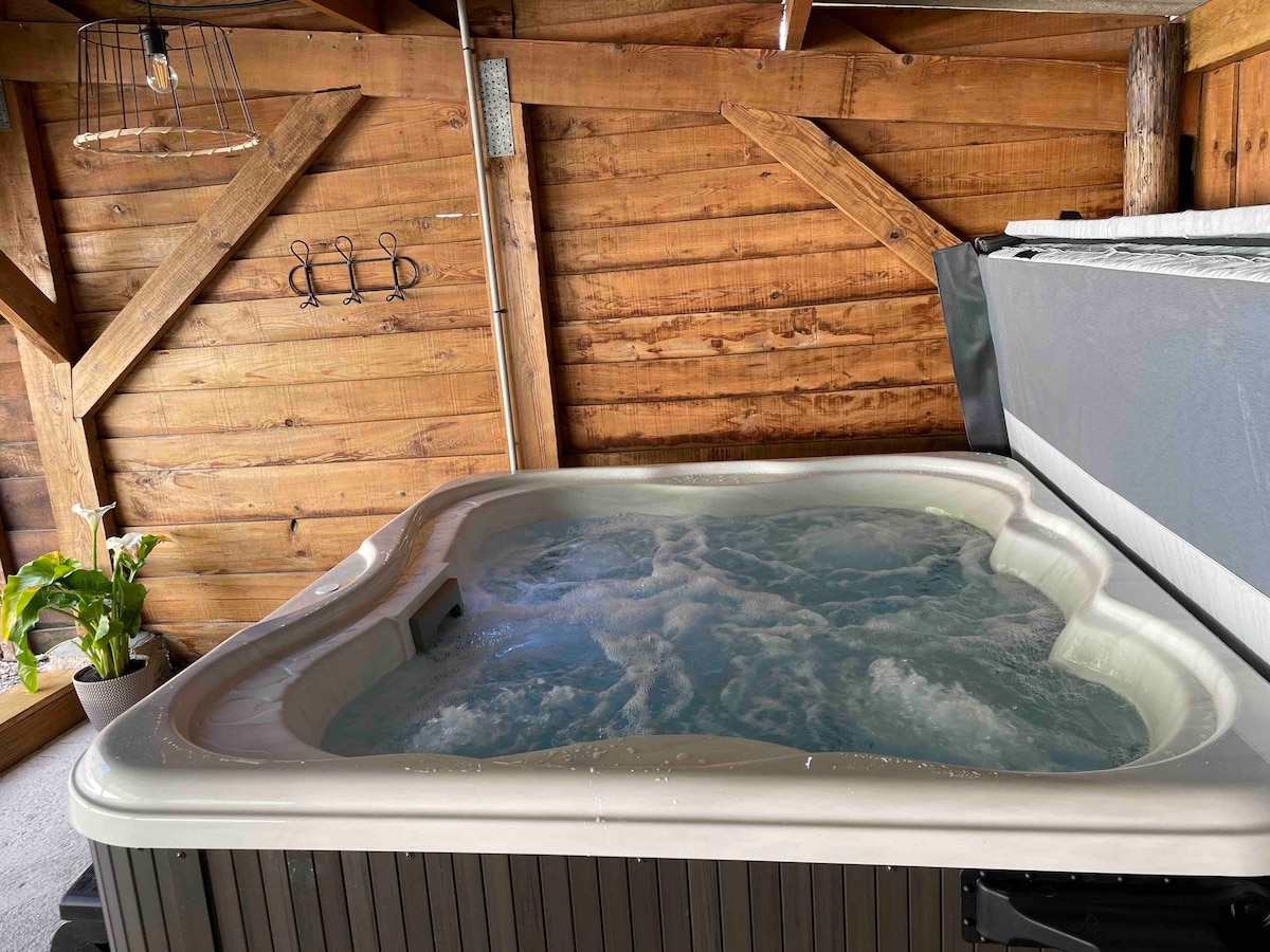 A hot tub is positioned in a wooden indoor space, surrounded by natural wood paneling. Gentle water movement is visible in the tub, and a potted plant adds a touch of greenery. Wall hooks are affixed above the tub for convenience.