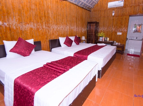 A sleeping area features two double beds, each dressed in white linens with patterned maroon throws. Accent pillows add a pop of color. The bamboo wall offers a natural texture, while a small table and lamp sit beside the beds, providing additional functionality.