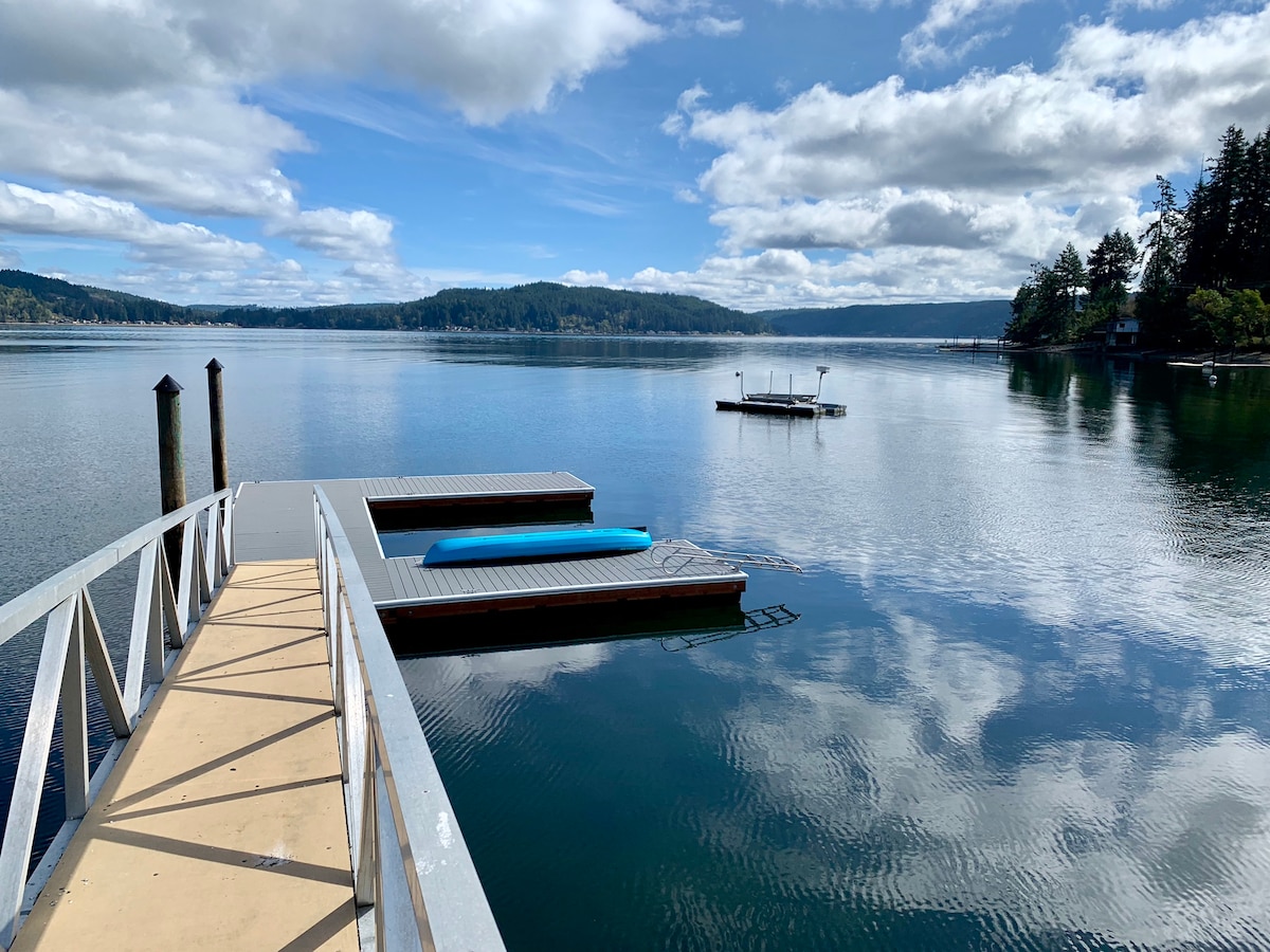 A calm waterfront scene featuring a dock extending over tranquil waters. A blue kayak rests on one of the floating platforms, while the mirror-like surface reflects the clouds and surrounding trees. The peaceful setting invites relaxation and exploration of the shoreline.