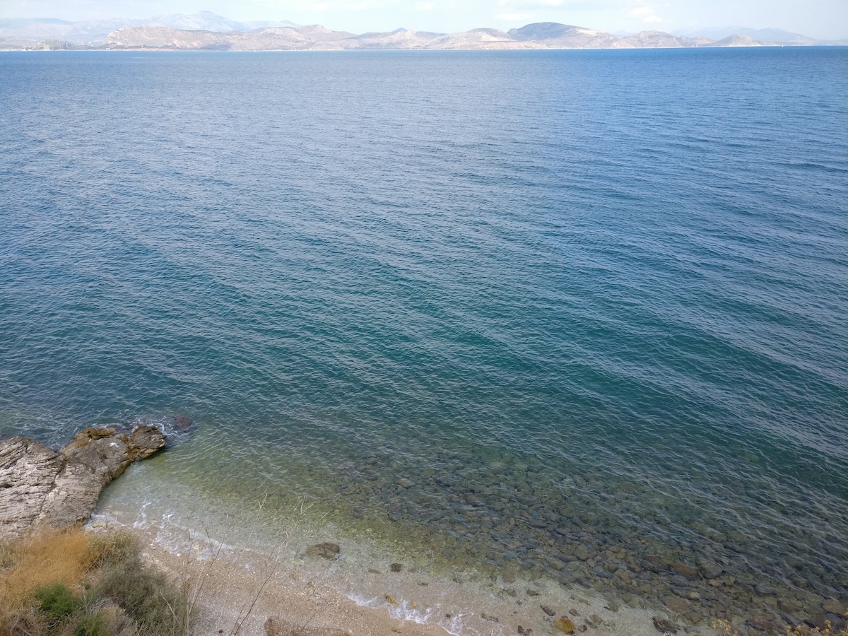 The image captures a serene view of the semi-rocky beach with clear, calm waters extending to the horizon. The shoreline features smooth pebbles and sandy areas, reflecting the natural beauty of the coastline, while gentle waves lap at the shore.