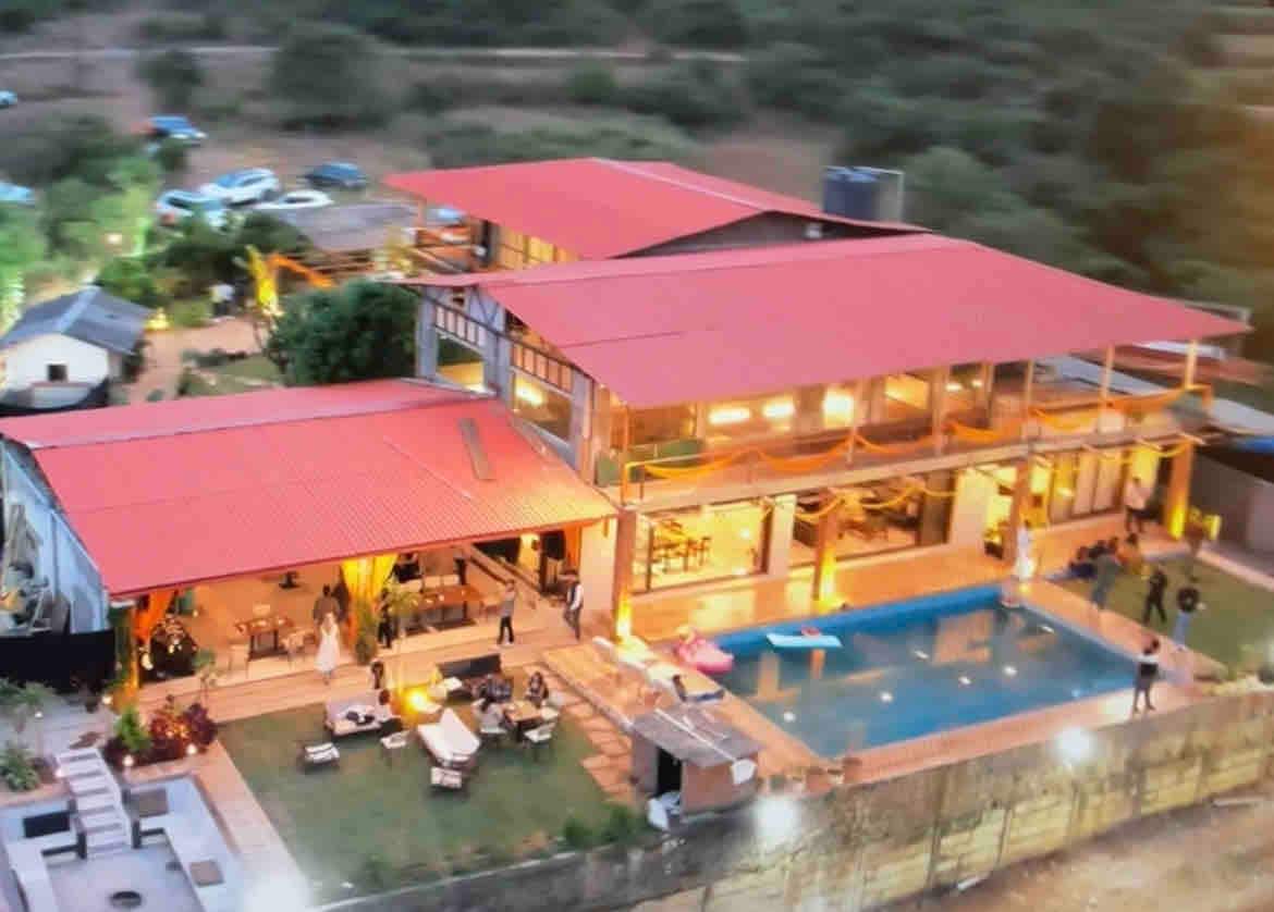 An aerial view of a luxurious vacation home showcases a red-roofed structure surrounded by greenery. A sparkling swimming pool is visible, with outdoor seating arranged nearby. The warm glow of lights illuminates the spacious outdoor area, enhancing the inviting atmosphere of this retreat.