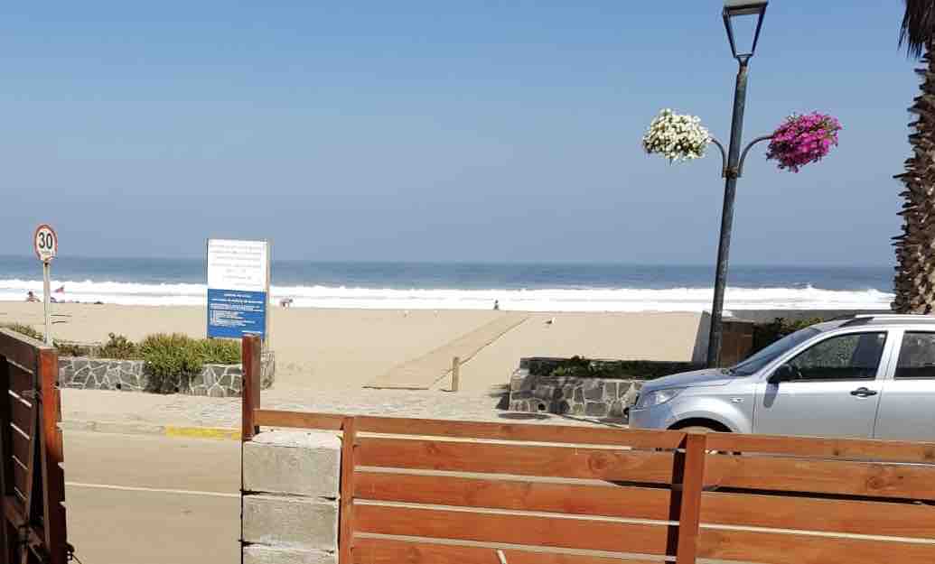A clear view of the beach is presented, showcasing soft sandy shores and gentle waves lapping at the coast. A road is visible in the foreground, bordered by a wooden fence and flowering planters. The scene is framed under a bright blue sky.