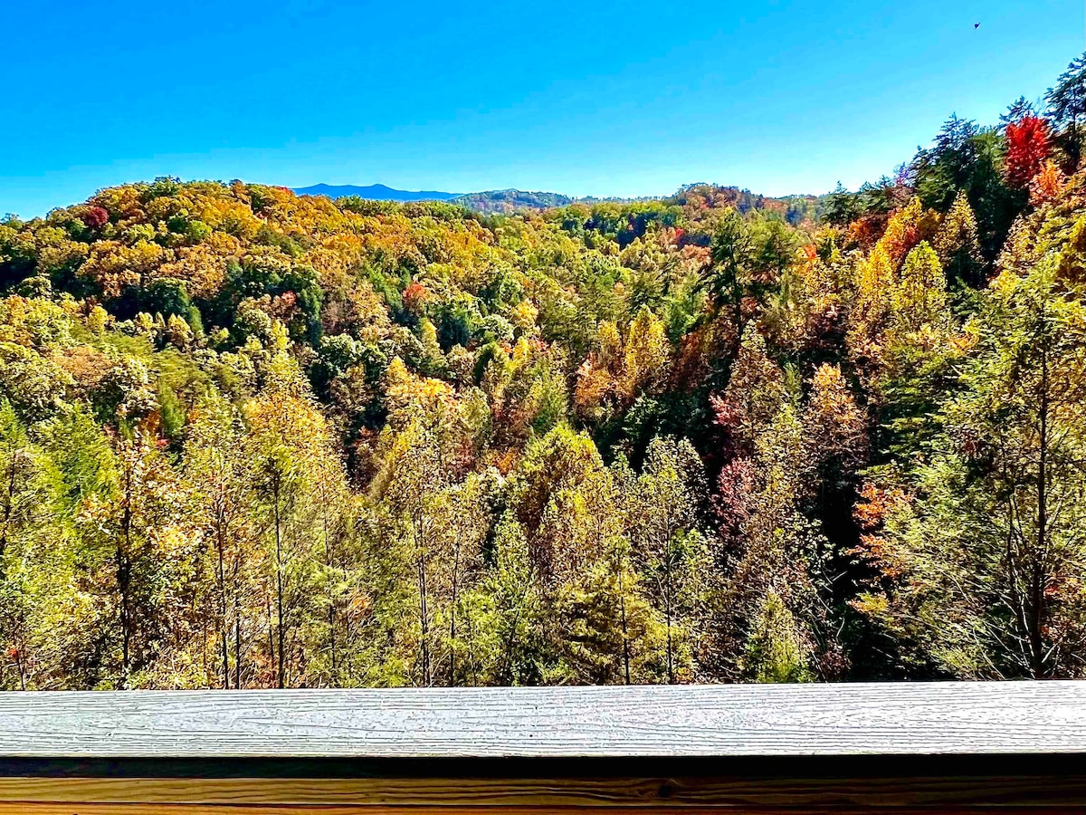 A panoramic view showcases a vibrant landscape of autumn foliage, with hues of orange, yellow, and green trees extending across rolling hills beneath a clear blue sky. The foreground features a wooden railing, framing the natural scenery.