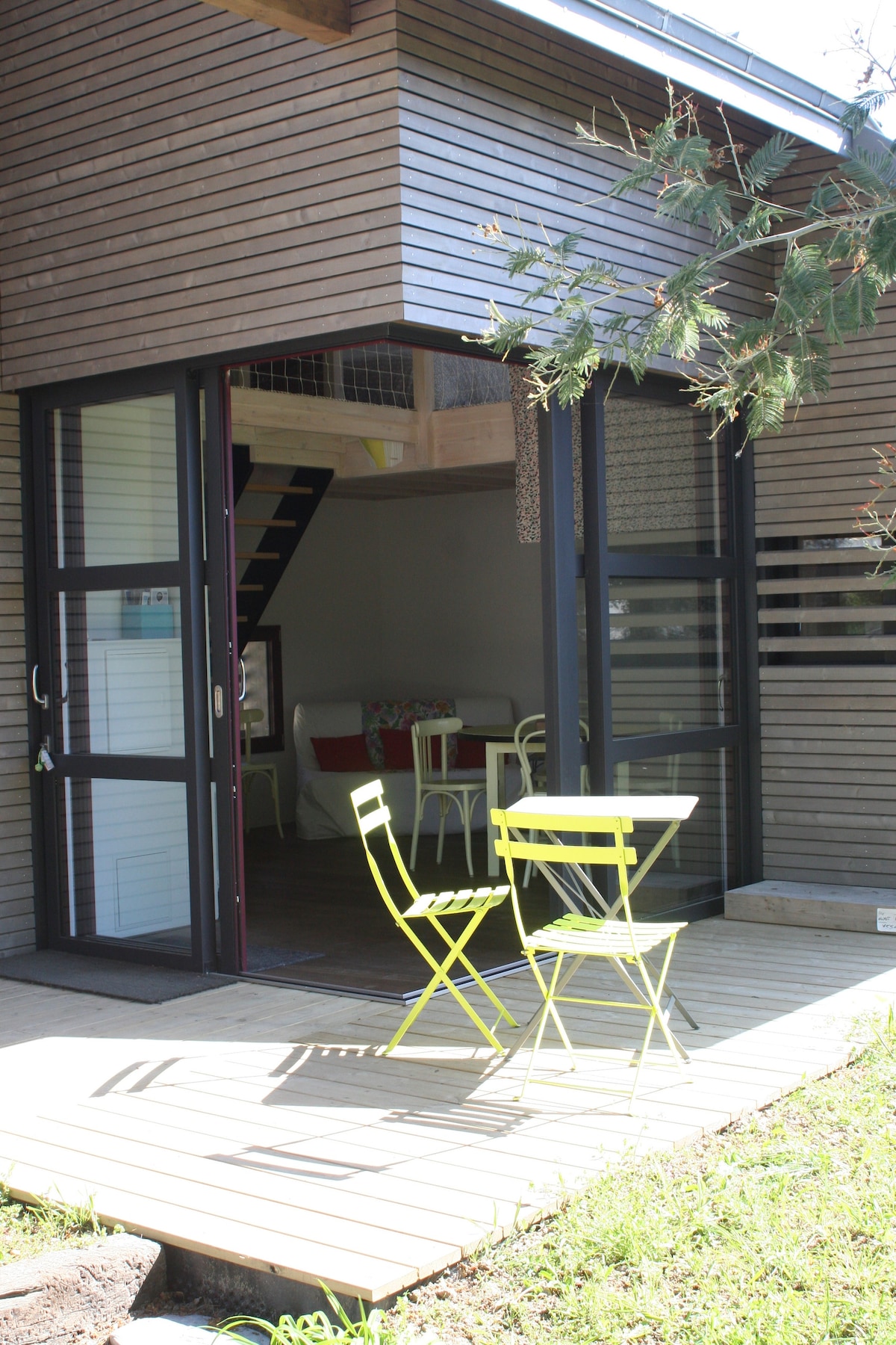 An outdoor terrace is shown, featuring two bright green chairs and a small table on a wooden deck. Large glass doors lead into a cozy interior space that is subtly visible, illuminated by natural light.