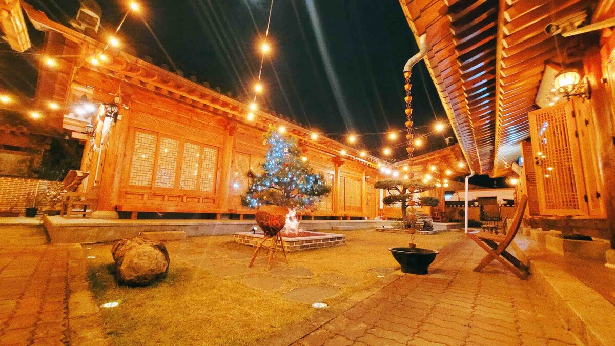 The outdoor space is illuminated by warm string lights, creating a cozy ambiance. A decorative tree is positioned in the center of the yard, surrounded by well-maintained grass and a few boulders. The traditional architectural elements of the hanok are visible on the sides.