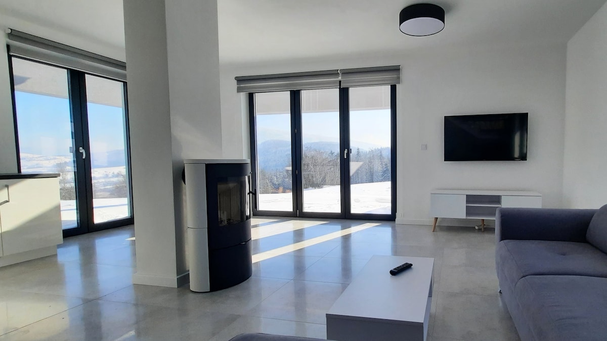 A bright living area features large glass doors that offer panoramic mountain views. A sleek fireplace contributes to the space, while a modern television is mounted on the wall. Light grey flooring and minimalistic furniture enhance the clean and open atmosphere.