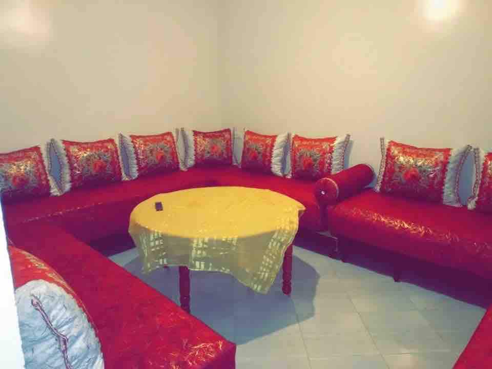 A cozy seating area features a large, red L-shaped couch adorned with decorative cushions. A circular yellow table sits in the center, providing a contrast to the vibrant seating. The walls are painted a light color, enhancing the room's brightness.