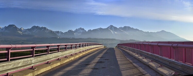 A curved bridge is seen extending towards a panoramic view of the High Tatras mountain range. Majestic peaks are outlined against a clear sky, with soft light creating a serene atmosphere. The bridge features a pink railing, framing the scenic landscape.