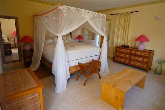 The bedroom features a large four-poster bed with a white canopy, complemented by pink bedside lamps. Natural light filters through the curtains, illuminating the light-colored walls. Two wooden storage pieces and a simple coffee table are arranged to enhance the room's spacious feel.