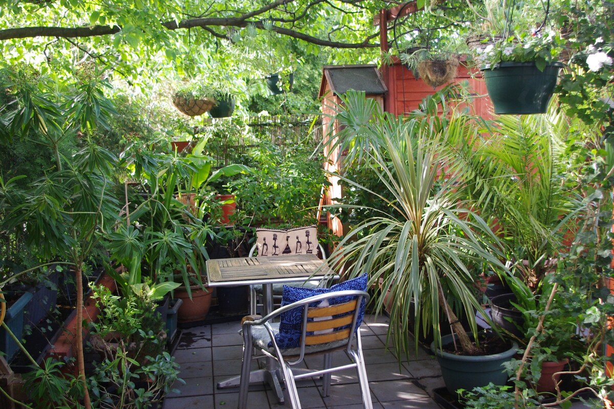 A lush terrace is surrounded by various potted plants and greenery, creating a verdant escape. A small table with chairs is positioned among the foliage, inviting relaxation in the serene outdoor space.