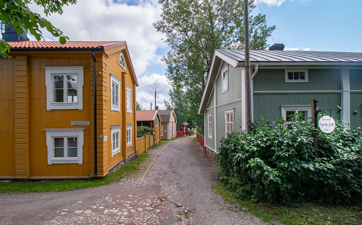 A narrow, tree-lined path is flanked by two charming buildings in warm colors. A yellow wooden house stands on the left, while a green house is on the right. Lush greenery borders the walkway, inviting exploration through this peaceful setting.