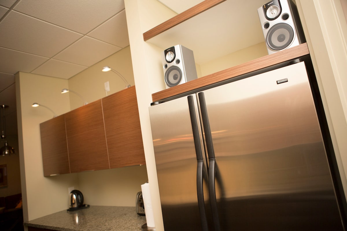 A modern kitchen is presented with stainless steel appliances, including a refrigerator and granite countertops. Under-cabinet lighting illuminates the space, highlighting wooden cabinetry and two speakers placed on top. A kettle is visible, contributing to the fully equipped cooking environment.