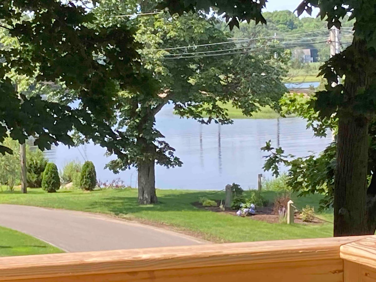 The image captures a serene view of a tranquil body of water bordered by lush green grass and mature trees. A quiet road runs adjacent to the scene, offering a peaceful natural setting. The reflection of the landscape is visible on the water's surface.