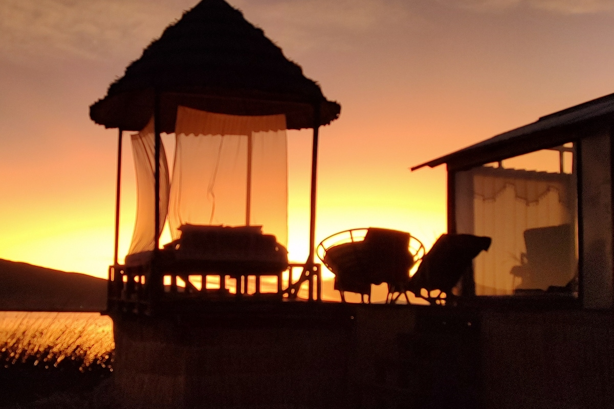 A serene outdoor seating area is framed by the silhouette of a thatched gazebo against a vibrant sunset. Comfortable chairs and a spacious bed are positioned to offer scenic views over the lake, enhancing the tranquil ambiance.