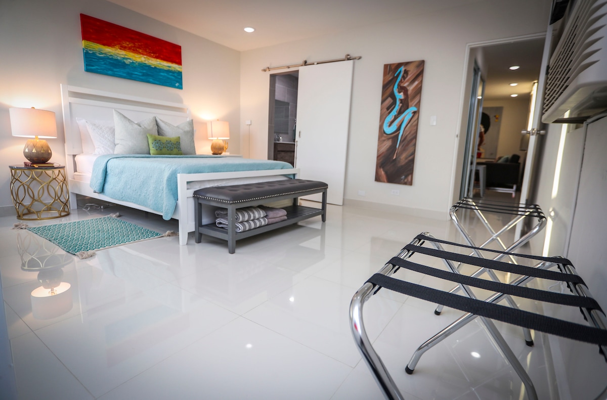 A stylish bedroom features a queen size bed with light bedding and decorative pillows. An abstract painting hangs above the bed, complemented by bedside tables with lamps. A storage bench sits at the foot of the bed, while a small rug adds a touch of color to the glossy floor.