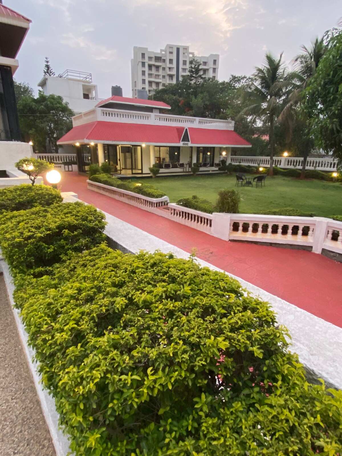 A well-manicured lawn surrounds a charming villa with a red roof. Pathways lead through the lush greenery, which is accented by soft outdoor lighting. The scene is set against a backdrop of nearby buildings and palm trees, creating a serene outdoor space.