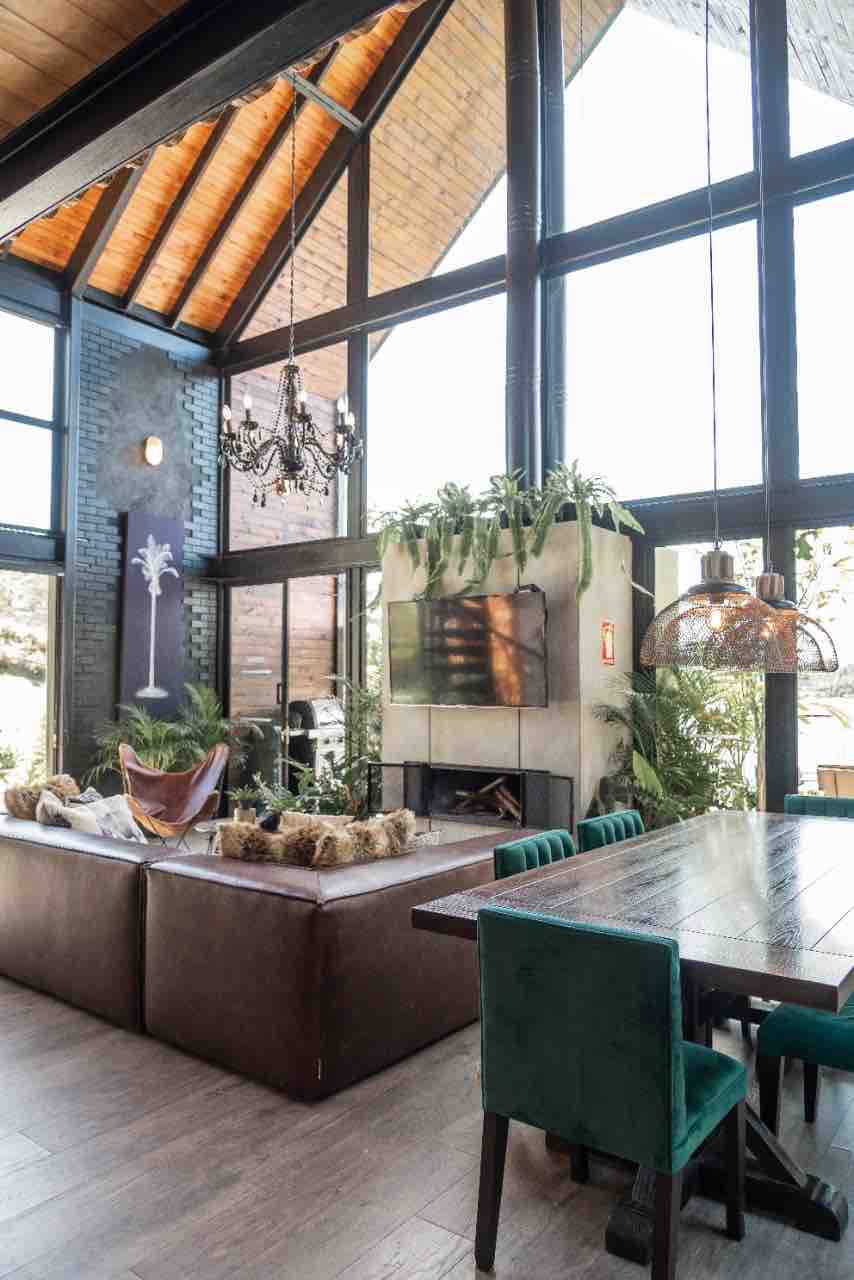 A spacious living area is characterized by high ceilings and large windows allowing natural light. A modern fireplace is set within a stylish wall, surrounded by greenery. A dining table is positioned adjacent to a comfortable lounge area furnished with a hammock.