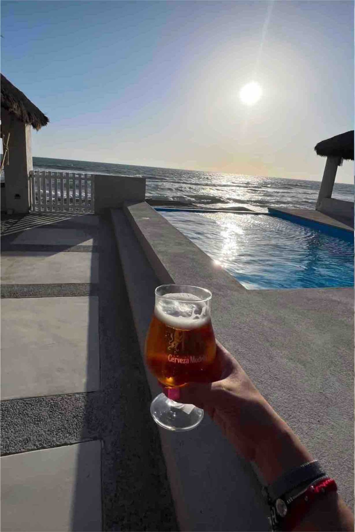 A glass of beer is held in front of an expansive ocean view, with the sun setting on the horizon. A shimmering pool is visible nearby, and the edge of a cozy shaded terrace adds to the serene atmosphere.