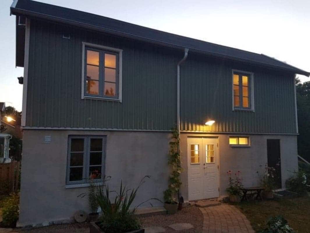 The exterior of a renovated house is shown during twilight, with lights illuminating the entrance. Windows on the upper and lower levels reflect a warm glow. The path is bordered by plants, contributing to a welcoming atmosphere.
