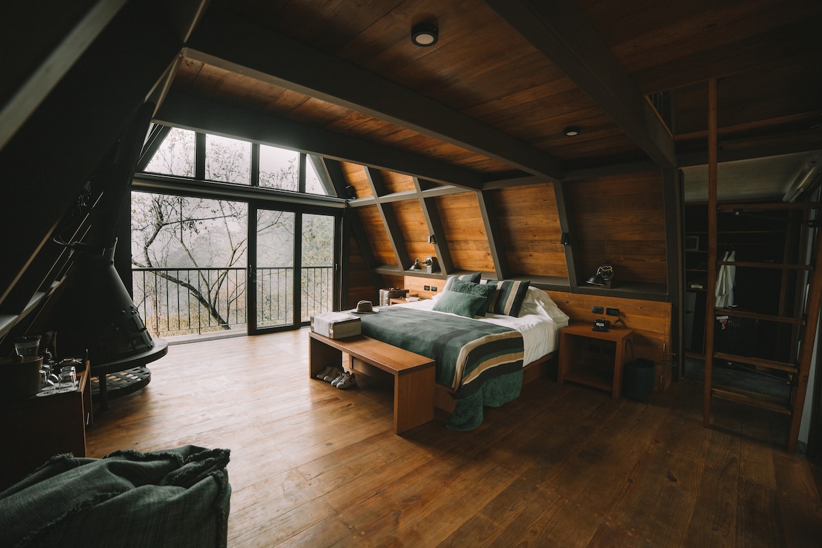A spacious bedroom features wooden walls and flooring, creating a warm ambiance. The bed is neatly arranged with soft linens, accompanied by a wooden table and chairs. Large windows offer natural light and views of the surrounding nature, while a modern fireplace adds a cozy touch.
