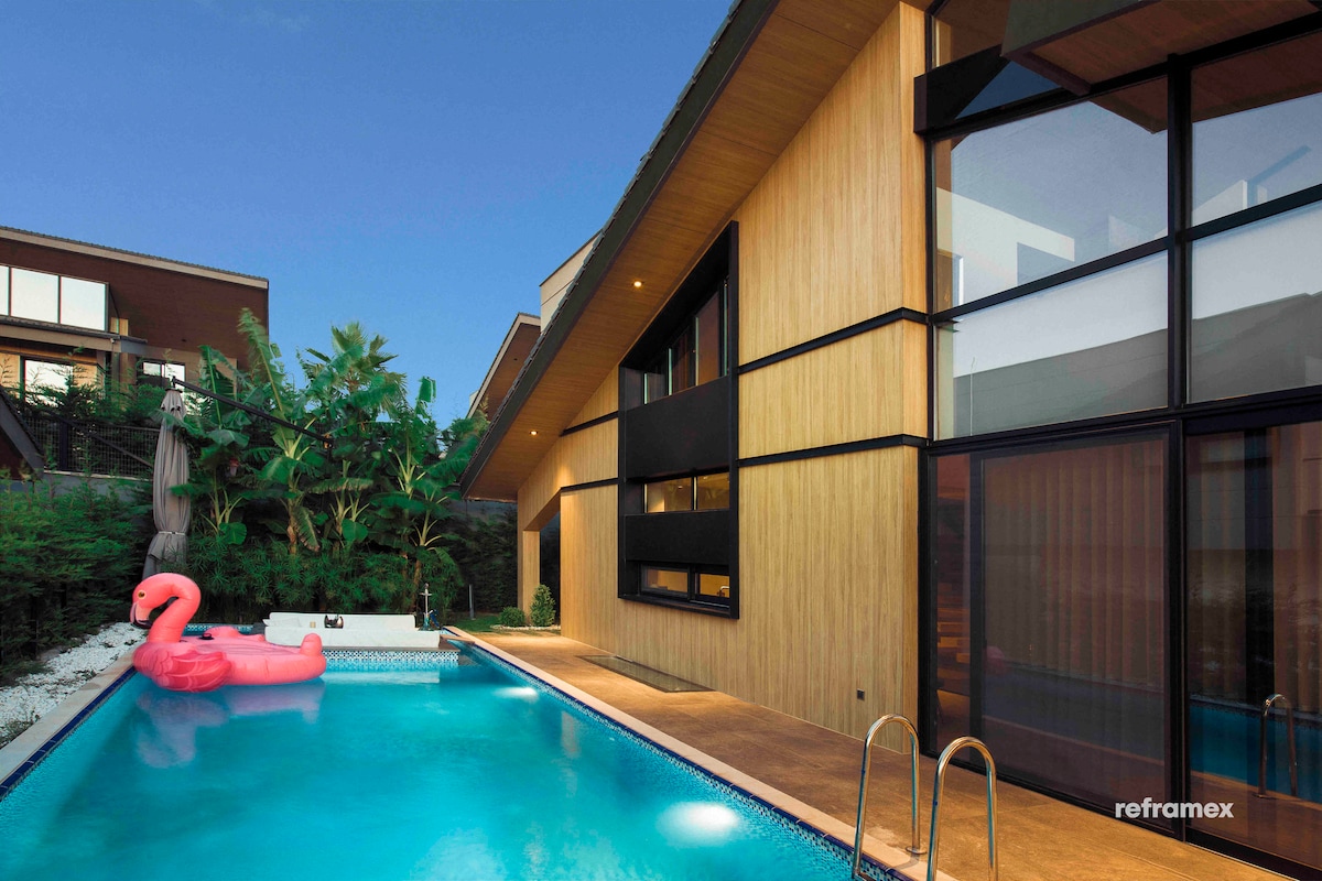 A modern villa features a heated pool surrounded by greenery. A light pink inflatable flamingo floats in the water. The exterior showcases a combination of wood paneling and large glass doors, inviting natural light into the space.
