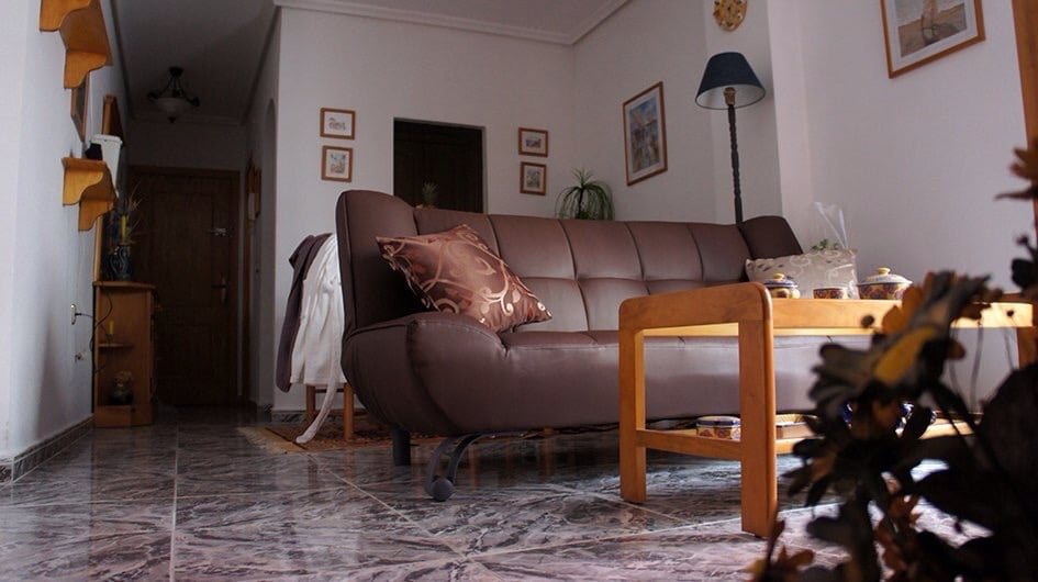 The living area features a brown, convertible sofa with decorative cushions. A small wooden coffee table is situated nearby on a tiled floor. The space is illuminated by a floor lamp, and wall art and indoor plants add a touch of decor.