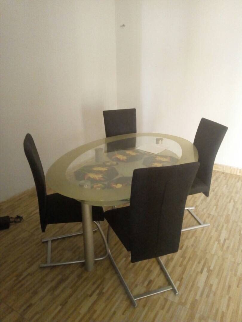 A round dining table with a glass top is surrounded by four high-backed black chairs. The table features a colorful decorative centerpiece. The light wooden floor complements the minimalistic design of the dining area.