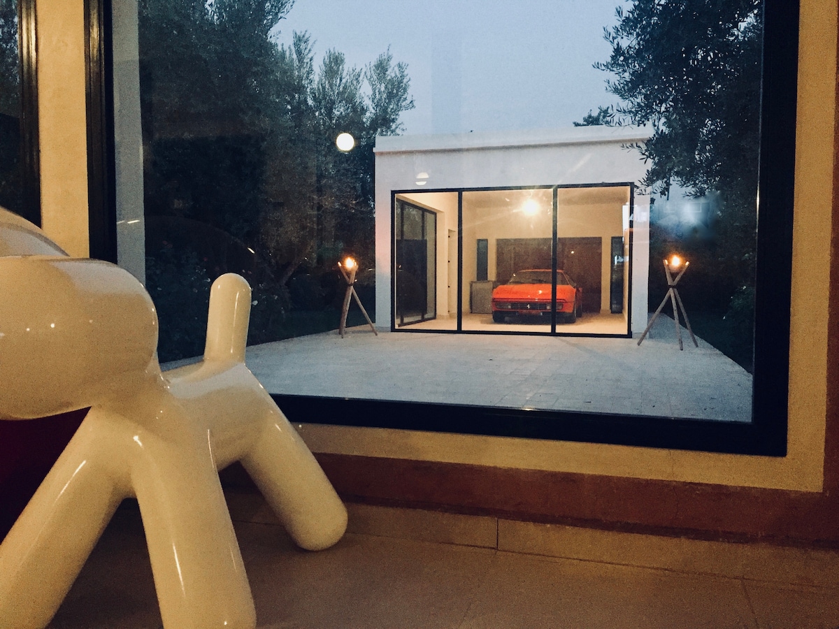 A view from inside the villa showcases an inviting outdoor space featuring a spacious patio. A red sofa is positioned under ambient lighting, flanked by decorative lamps. The modern structure is framed by large glass doors, blending the indoor and outdoor environments.