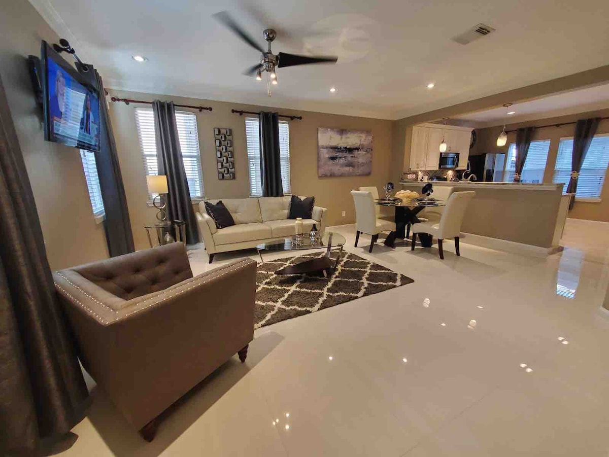 A contemporary living area is presented with a neutral color palette and modern décor. A comfortable sofa and armchair are arranged around a glass coffee table, while large windows offer natural light. A dining area with a round table is visible in the background.
