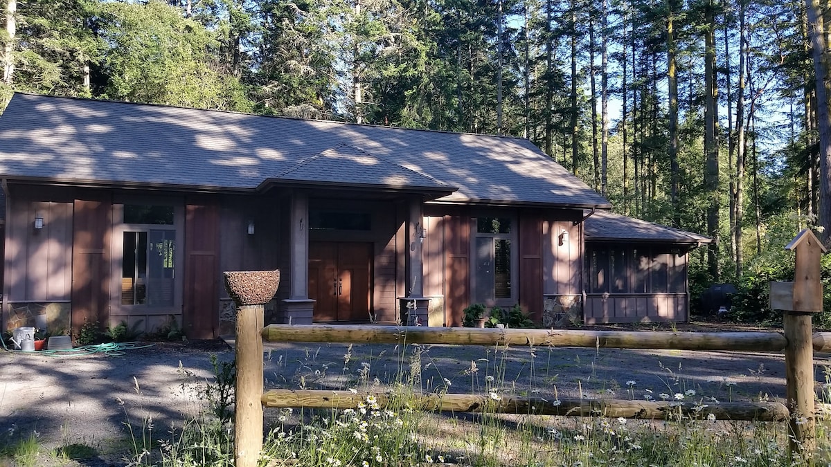 A single-story structure is set within a wooded area, featuring a combination of large windows and a wooden façade. A wooden fence surrounds the front yard, while various plants and wildflowers are visible in the foreground, enhancing the natural setting.