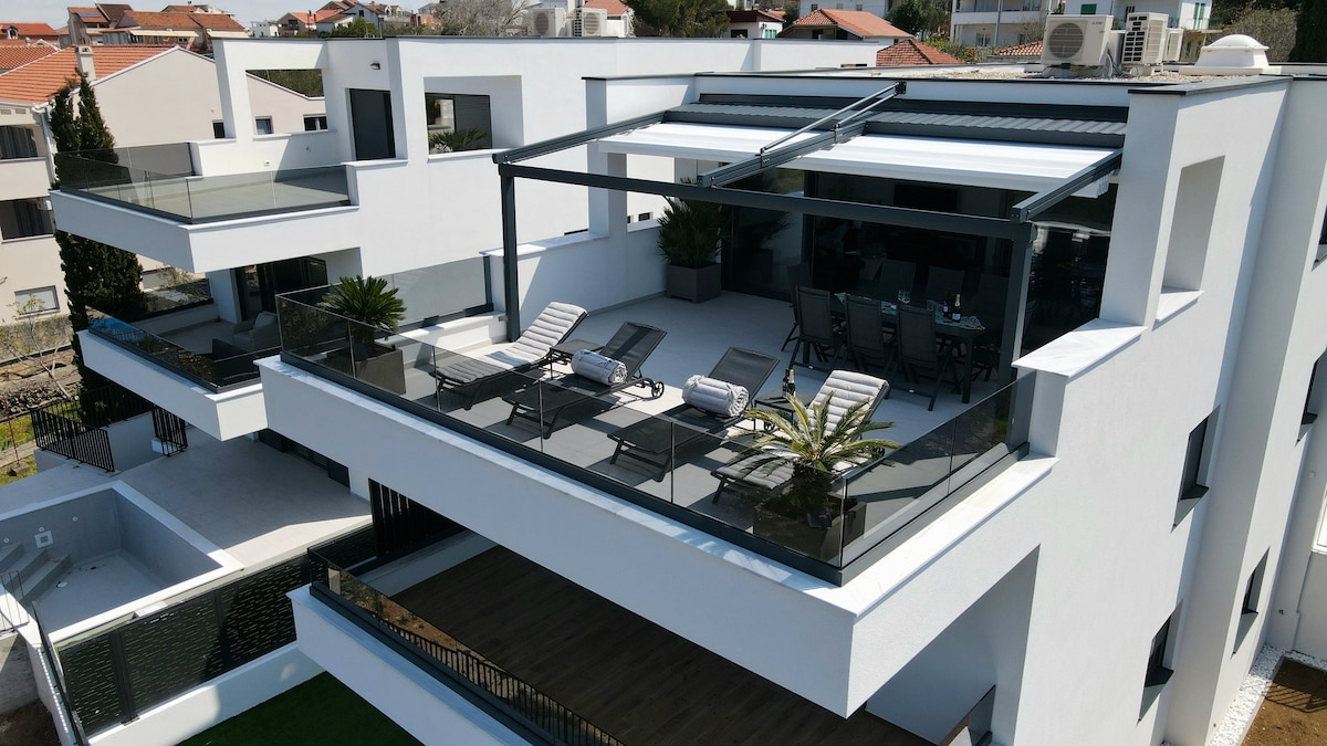 An elevated terrace features several lounge chairs and a dining area, surrounded by glass railings. Potted plants are placed strategically, enhancing the outdoor space. The modern architecture presents clean lines and ample natural light.