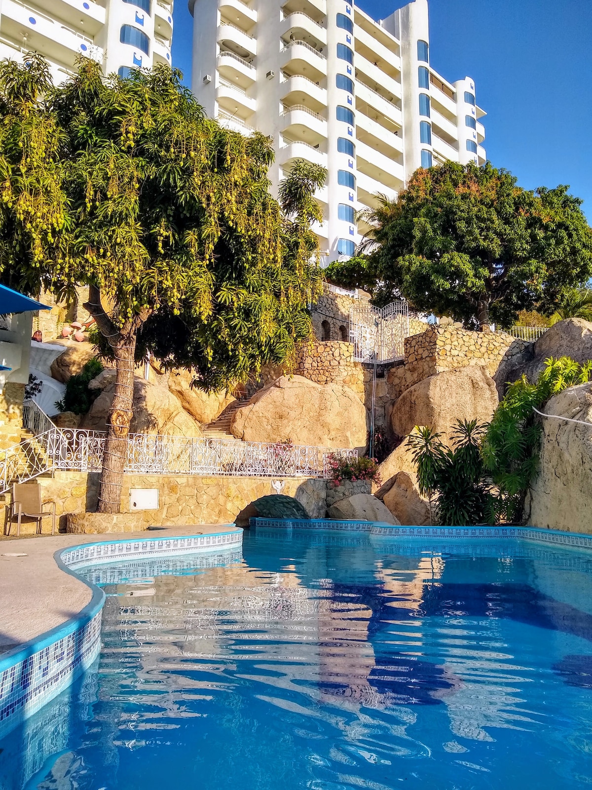 A calm pool is surrounded by natural rock formations and lush greenery. Water cascades gently from the rocks, adding a serene touch to the scene. The structures of a multi-story building rise in the background under a clear blue sky.