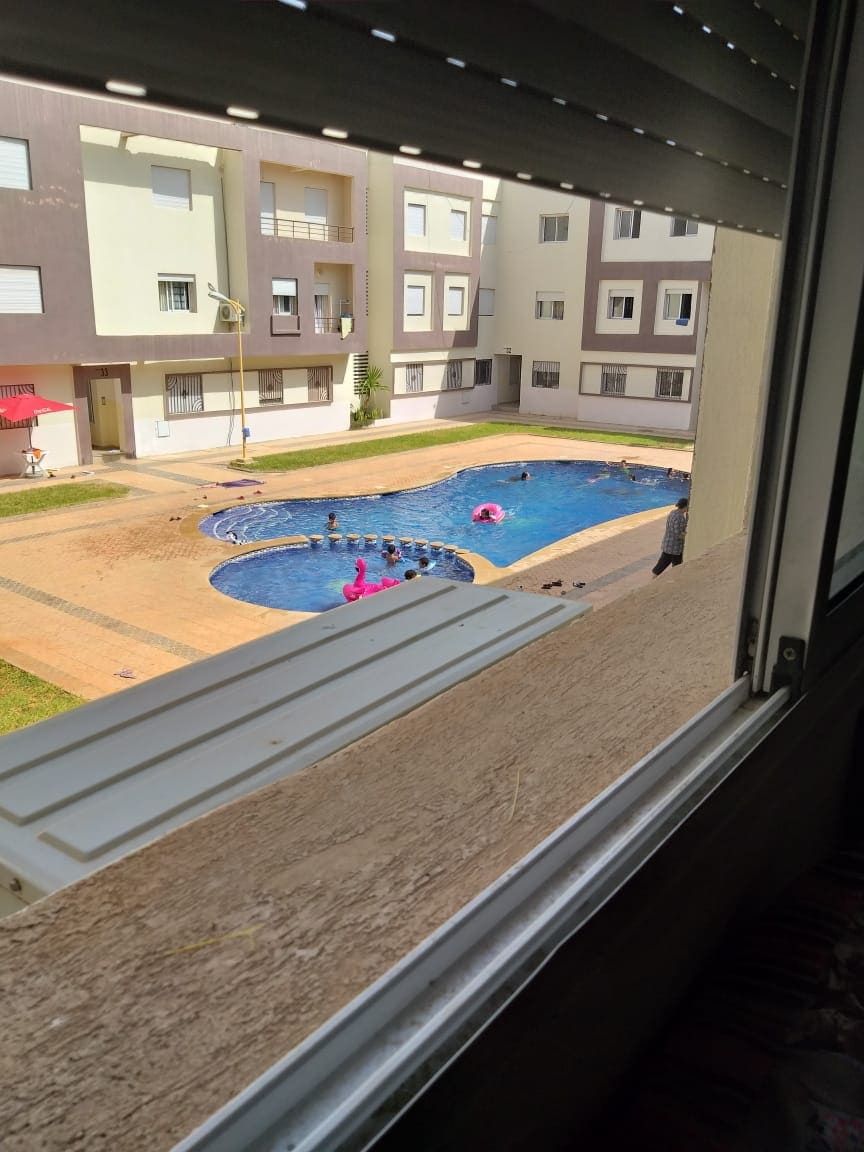 A view from a window captures a communal pool surrounded by a landscaped area. Swimmers can be seen enjoying the water, with colorful inflatables adding vibrancy. The modern building architecture and patio area enhance the overall ambiance of the space.