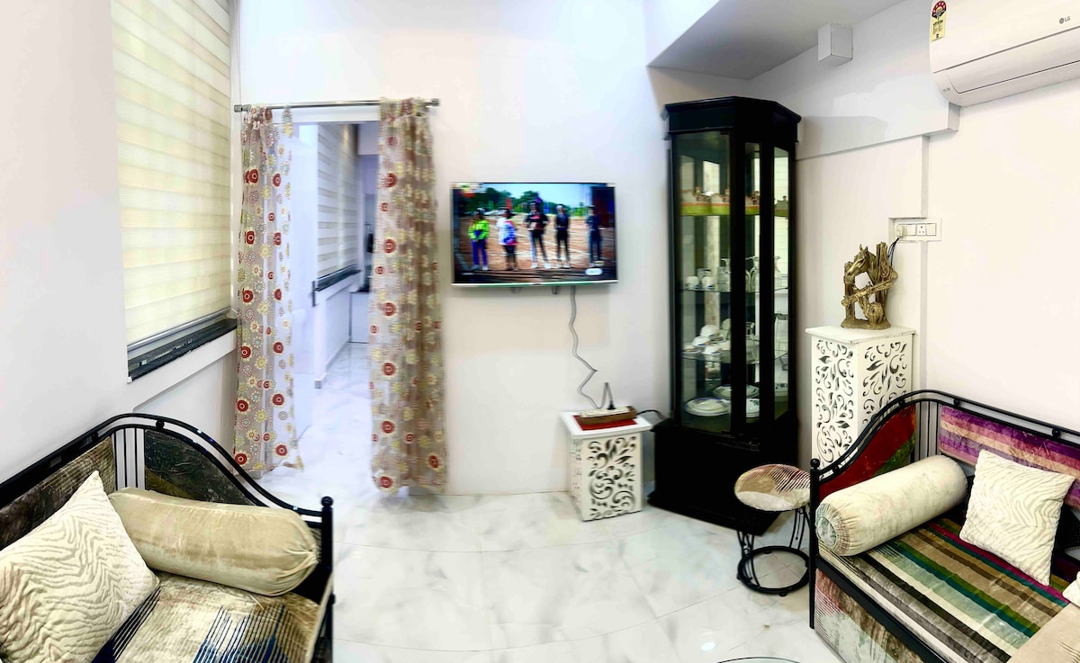 A well-lit living space features a television mounted on the wall and a glass cabinet displaying decorative items. Comfortable seating is arranged, including a two-seater and a single couch, with patterned curtains framing the window. Light-colored marble flooring contributes to the bright atmosphere.