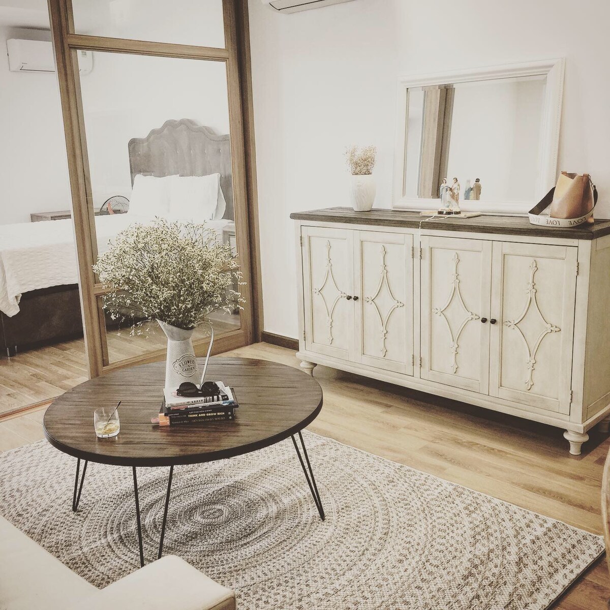 The living area is furnished with a low round coffee table positioned on a textured area rug. A stylish sideboard with decorative features stands against the wall, while a mirror reflects the space. Fresh flowers are placed in a vase atop the table, adding a touch of warmth.