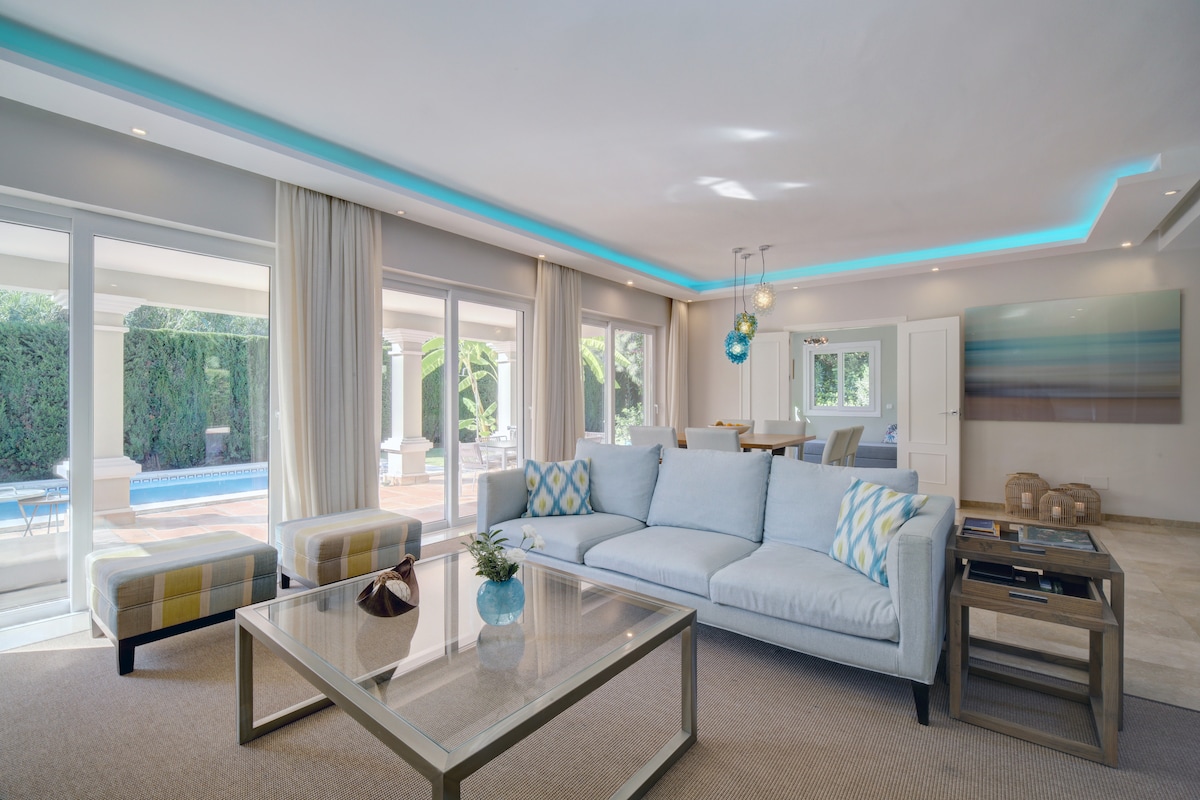 A spacious living area features light-colored furniture, complemented by decorative pillows. Large glass doors provide views of the private pool area, allowing natural light to fill the room. An elegant dining space is visible in the background, alongside modern light fixtures that enhance the ambiance.