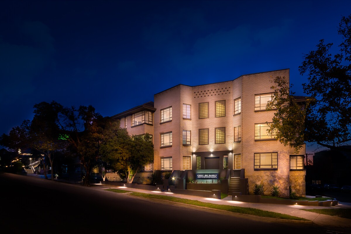 The building's exterior is illuminated at night, showcasing its modern architectural design. Soft lighting highlights the entrance and landscaping, while numerous windows reflect warm tones. A peaceful street is visible in front, enhancing the welcoming ambiance of the property.
