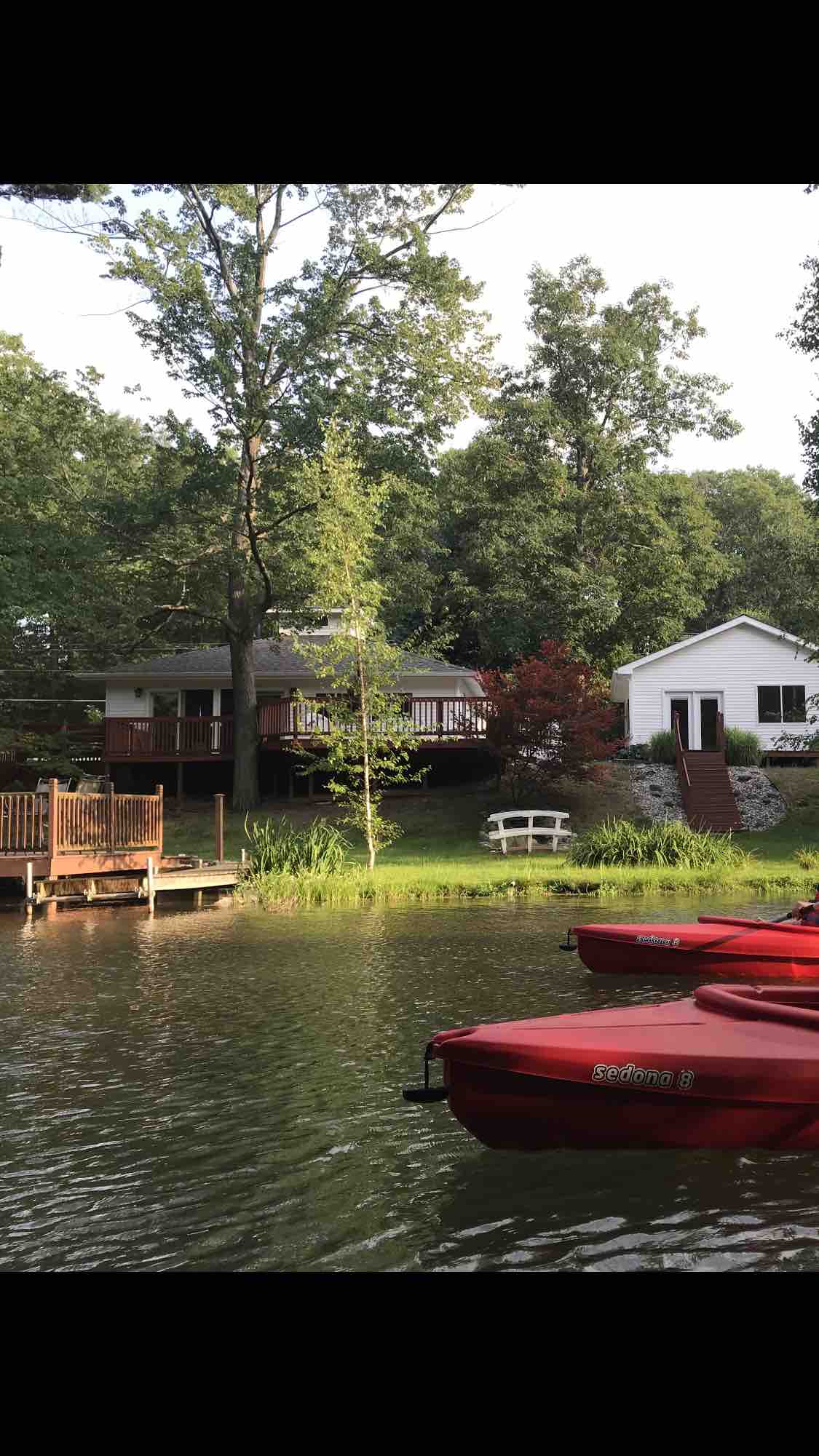 Red paddle boats are anchored near the shoreline, surrounded by lush greenery. A large deck is visible, leading to a cozy cottage nestled among trees. The property features a peaceful waterfront setting with a wooden bench positioned beside the water.