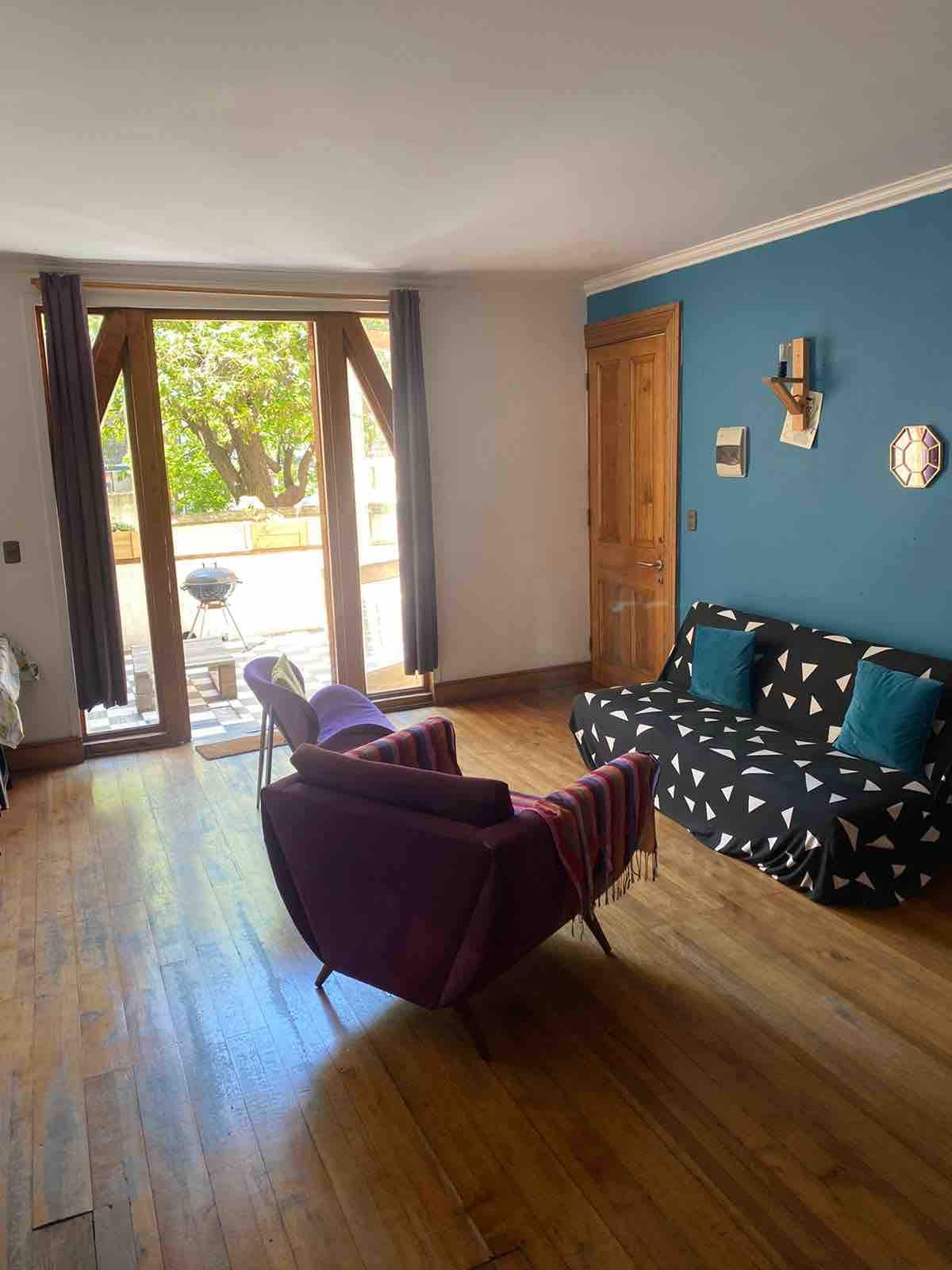 A cozy living area is featured with dark wooden floors and a rich blue accent wall. Two chairs are positioned next to a small couch adorned with a geometric pattern. Large glass doors open to a terrace, allowing natural light to brighten the space.