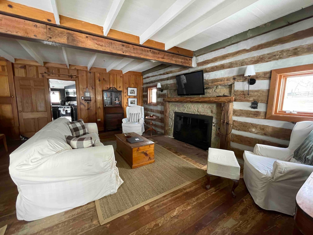 A cozy log living room is showcased with rustic wooden beams and walls. The space features a fireplace and a television. Two white slipcovered chairs and a couch are arranged around a wooden coffee table, with a woven rug adding warmth underfoot.