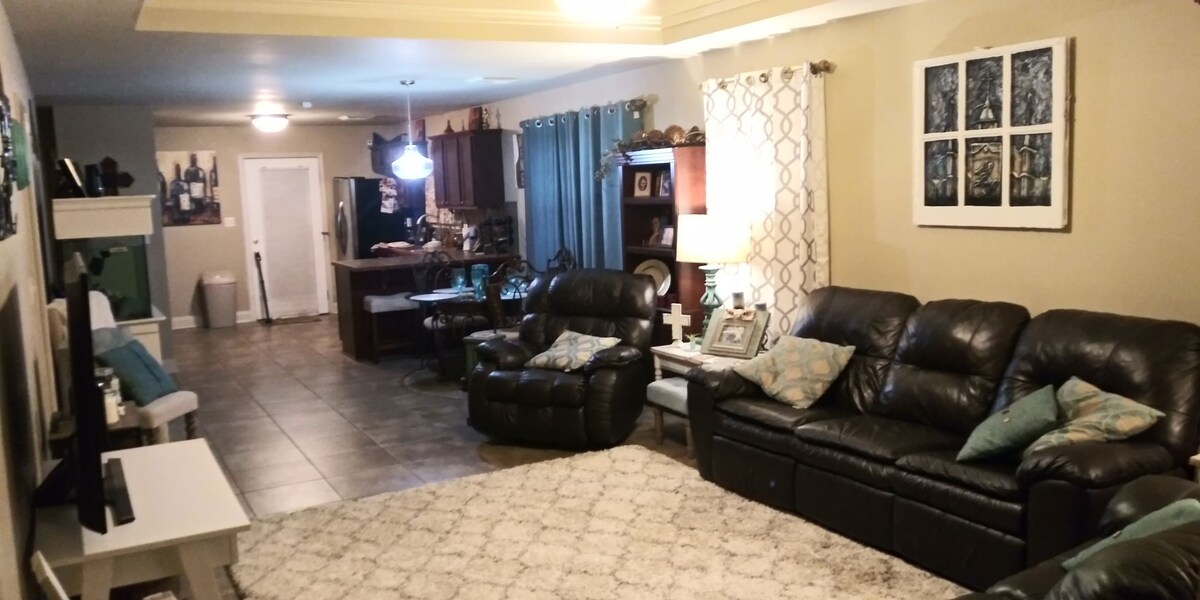 The living area features comfortable leather recliners arranged around a light rug, enhancing the inviting space. Shelves hold decorative items, while art is displayed on the walls. Natural light enters through large windows with light curtains, leading to an open kitchen area in the background.