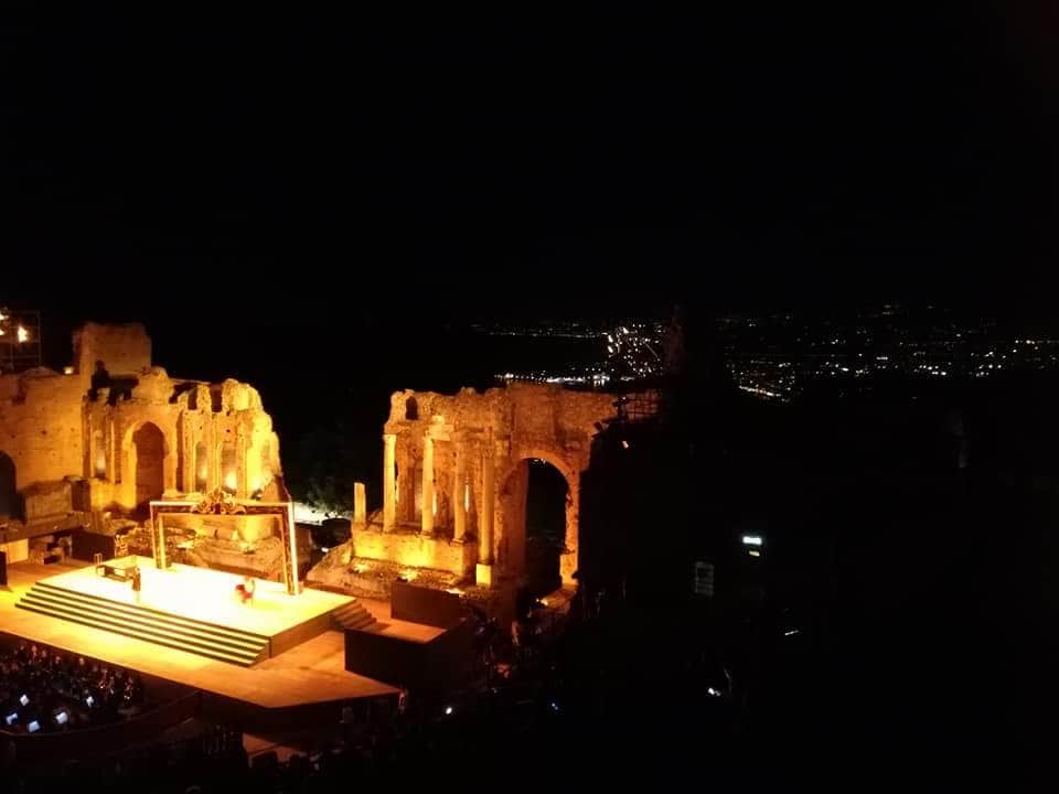 An atmospheric view of an ancient theater illuminated at night, showcasing the warm glow of the stone ruins. The stage is set for a performance, while distant city lights twinkle in the background, adding to the enchanting scene.