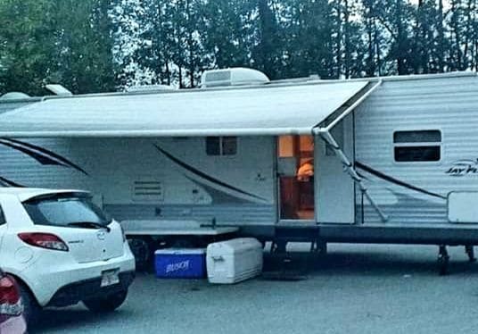 A travel trailer is parked in a natural setting, showcasing its exterior with an awning extended. A nearby vehicle is visible, along with a cooler positioned in front of the trailer. The surroundings include leafy trees, suggesting a retreat into nature.