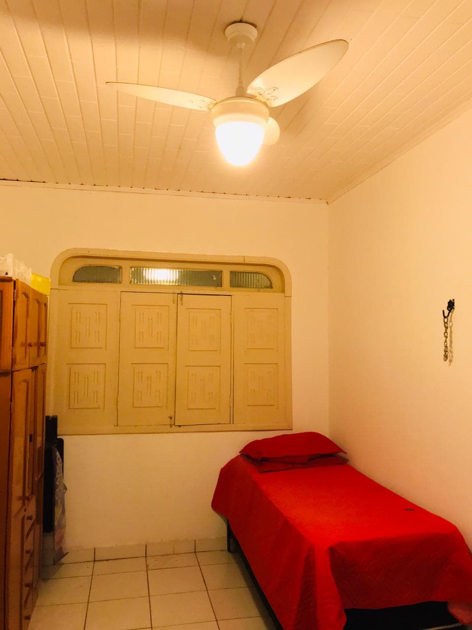 A small room features a cozy single bed with a red coverlet, positioned against a light-colored wall. A ceiling fan provides air circulation, while a window with paneled shutters allows natural light to enter. A wooden wardrobe is visible, offering storage space.