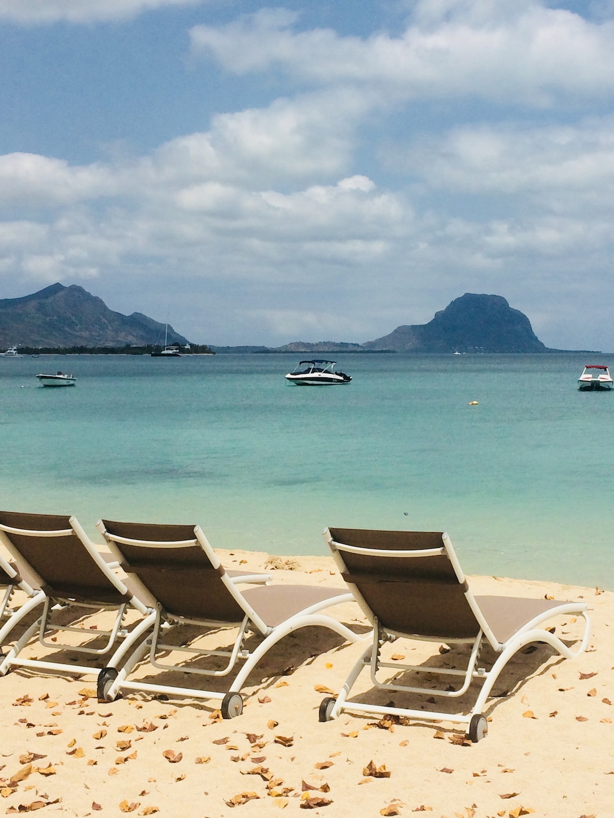 Several lounge chairs are arranged on the sandy beach, facing the calm, turquoise sea. The distant Le Morne mountain is visible on the horizon, alongside a few boats floating gently in the water under a partly cloudy sky.